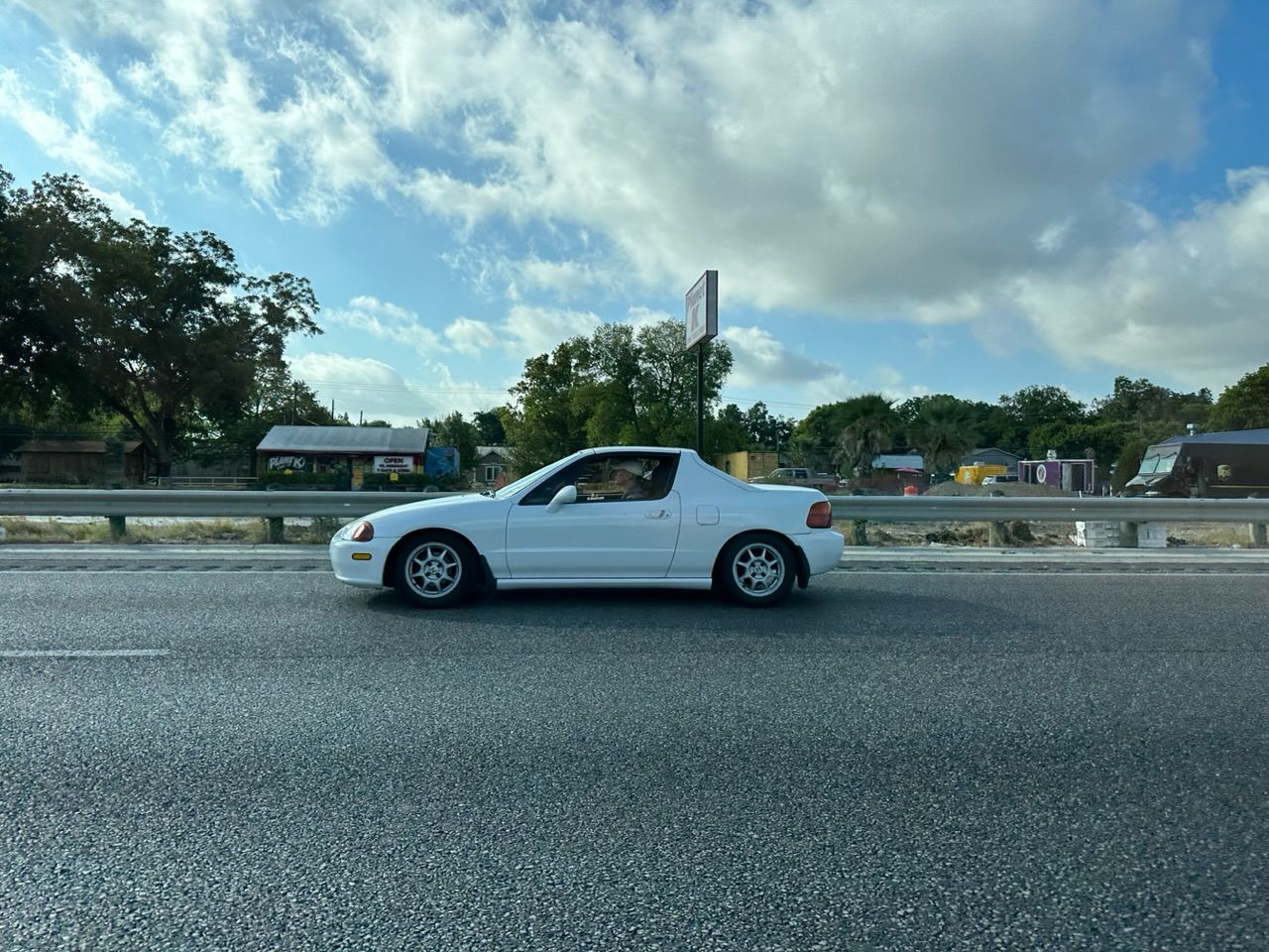 A white Del Sol, rolling along. The driver looks focused, despite a drink in his hand.