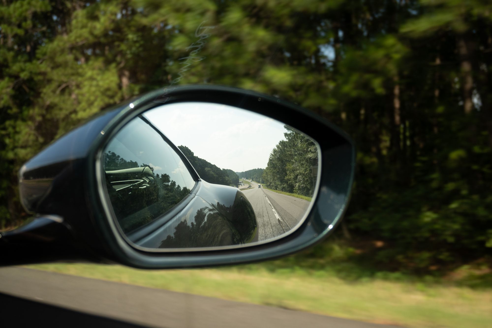A photo in the passenger side mirror of the car. Trees slip by in the background, while the mirror shows the rear fender of the car and the road behind. There’s a mild hill and curve in the road, the grass is green, and trees line both sides of the freeway.