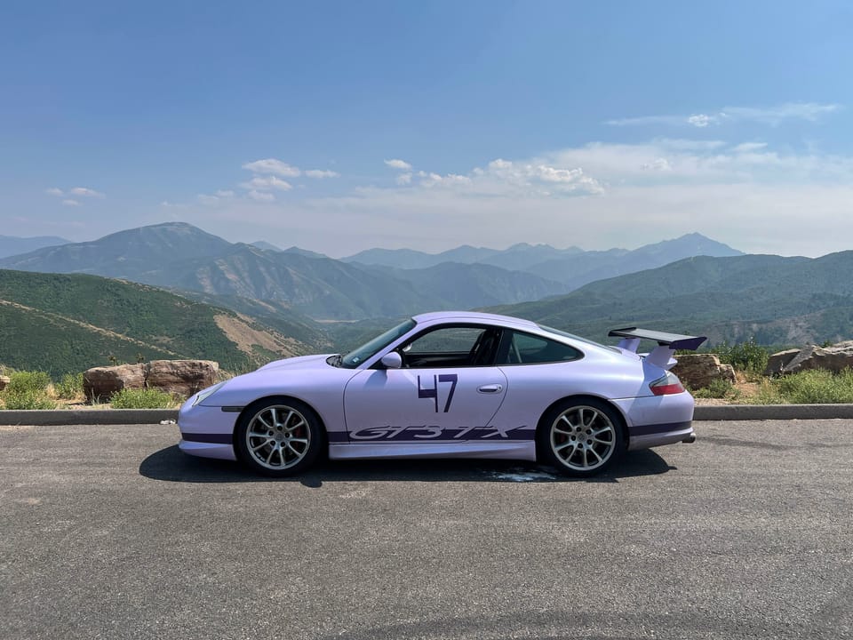 Lavender and Purple Porsche GT3 with race numbers sports car parked on the side of the road with a scenic mountain background